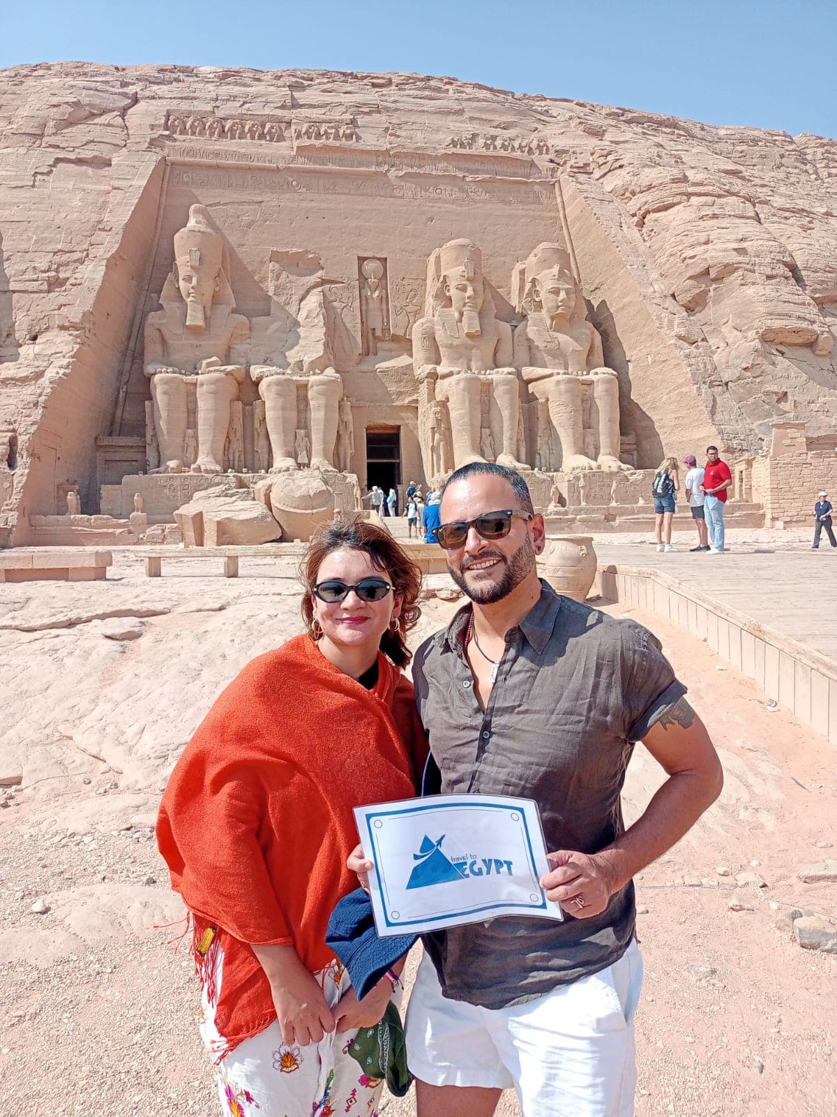 Couple holding the Travel To Egypt sign in front of the colossi of Ramesses II at Abu Simbel